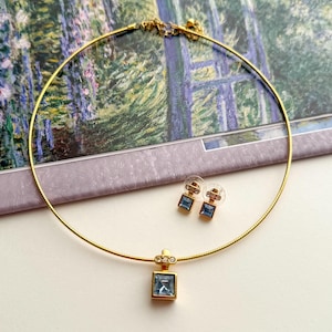 May include: A gold-toned necklace with a square, light blue pendant and matching stud earrings. The necklace and earrings feature a gold setting with clear accent stones. The jewelry set is displayed on a light-colored surface.