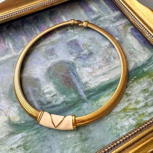 Può includere: Una collana girocollo dorata con un design a coste e un elemento centrale color crema. La collana è esposta su un dipinto all'interno di una cornice dorata ornata. Lo sfondo presenta uno stile morbido e impressionistico.