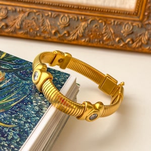 May include: A gold-toned metal bracelet featuring a flexible, ribbed band design. The bracelet is accented with two smooth, heart-shaped gold elements, each set with a single clear, round-cut gemstone in the center, and includes a rectangular clasp.