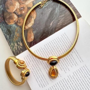 May include: A gold-toned necklace and bracelet set. The necklace is a flexible, ribbed design with a pendant featuring a black and amber-colored stone. The bracelet has matching stones. The jewelry is displayed on a printed page.