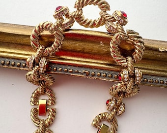 Könnte beinhalten: Ein goldfarbenes Armband mit einem gedrehten Seildesign. Das Armband verfügt über rote und klare Edelsteine und einen Verschluss. Der Schmuck wird auf einer goldgerahmten Oberfläche präsentiert.