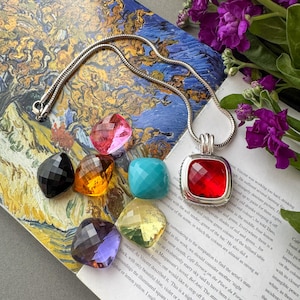 May include: A silver-toned snake chain necklace with a square pendant featuring a faceted red stone. Surrounding the pendant are six loose, faceted, cushion-cut stones in pink, black, amber, turquoise, purple, and yellow, all arranged on a printed page.