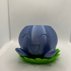 Può includere: Un vaso per piante stampato in 3D di colore blu scuro a forma di personaggio sorridente, appoggiato su una base verde a forma di foglia. Il personaggio ha un sorriso intagliato e braccia piccole.