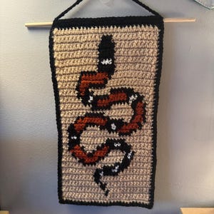 May include: Crocheted wall hanging with a snake motif. The rectangular piece has a beige background with a black border. The snake is in black, red, and white. A wooden dowel and black string are used for hanging.