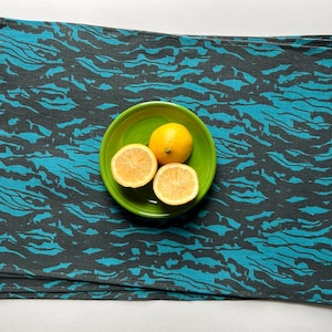 Puede incluir: Juego de cuatro manteles individuales de lino azul turquesa y negro. Los manteles individuales están apilados uno encima del otro. Un tazón verde con tres limones está encima de los manteles individuales.