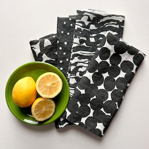 May include: A set of five black and white patterned kitchen towels. The towels are folded and arranged in a fan shape. Three lemons are on a green plate in front of the towels.