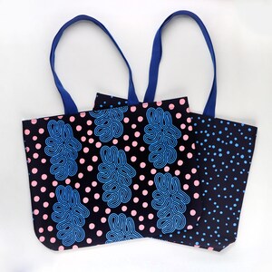 Puede incluir: Dos bolsas de mano con asas azules. Una bolsa tiene un fondo negro con lunares rosas y formas abstractas azules. La otra bolsa tiene un fondo negro con lunares azules. Las bolsas son de tela.
