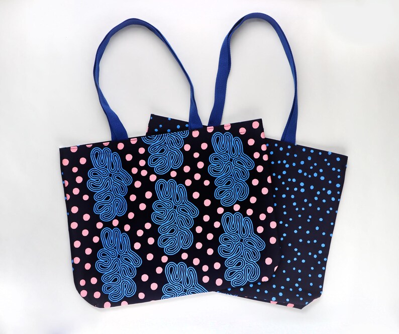 Puede incluir: Dos bolsas de mano con asas azules. Una bolsa tiene un fondo negro con lunares rosas y formas abstractas azules. La otra bolsa tiene un fondo negro con lunares azules. Las bolsas son de tela.