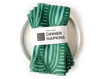 Pode incluir: Um conjunto de guardanapos de linho verdes e turquesas com um padrão abstrato repetido. São presos por uma faixa branca com a inscrição "Dinner Napkins" e "100% Linen". Colocados sobre um prato branco.