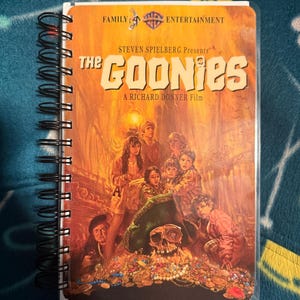 May include: A spiral-bound notebook featuring the movie poster for The Goonies on the front cover. The cover displays the film title, cast members, a skull, and a pile of treasure in warm orange and yellow tones with a black wire binding on the left.