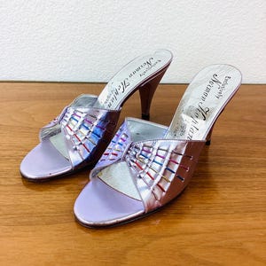 May include: A pair of lavender satin heeled sandals with a colorful woven detail on the straps. The sandals are labeled "Exclusively  Helman  New York".
