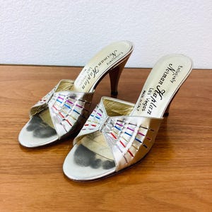 May include: A pair of silver metallic heeled sandals with a colorful woven strap design. The sandals are labeled "Norman Kaplan Exclusively Las Vegas Made in Italy".