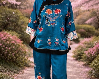 May include: Teal silk pajama set with floral embroidery. The top has a mandarin collar and black trim. The pants are wide-legged. The embroidery features flowers and a butterfly. The sleeves have a white and floral patterned cuff.