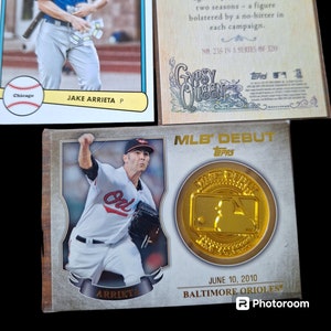 May include: Three baseball trading cards featuring Jake Arrieta, a pitcher for the Chicago Cubs and Baltimore Orioles. The cards are from the Topps Gypsy Queen and MLB Debut series. The cards feature photos of Arrieta pitching and text about his career.
