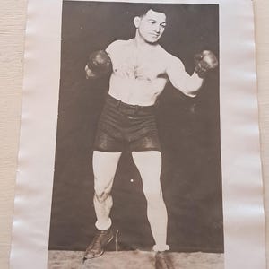 May include: Black and white vintage photograph of a boxer in a fighting stance. The boxer is shirtless, wearing dark shorts, boxing gloves, and boots. The image is a full-body shot against a dark background.
