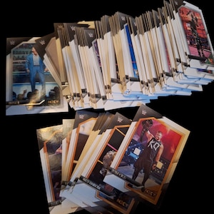 May include: A collection of trading cards featuring wrestlers, with action shots and the NXT logo. The cards are stacked and fanned out, with a black background. Some cards show wrestlers in action, with the NXT logo.