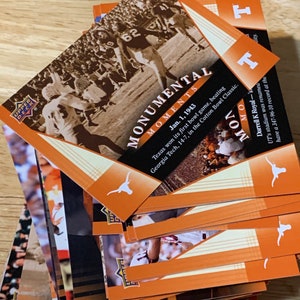 May include: A stack of orange and white trading cards featuring a Texas Longhorns football team logo and the words "Monumental Moments". The top card shows a black and white photo of a football game with the text "Jan. 1, 1943 Texas won its first bowl game, beating Georgia Tech, 14-7, in the Cotton Bowl Classic."