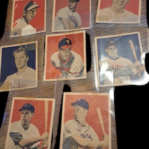 May include: A collection of eight vintage baseball cards featuring different players in their uniforms. The cards are in various conditions, some with minor wear and tear. The cards are displayed on a wooden surface.