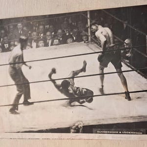 May include: Vintage black and white photograph of a boxing match in progress. One boxer is falling in the ring, while another stands ready. Spectators are visible in the background, watching the fight.
