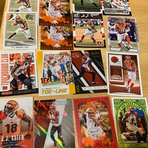 May include: A collection of 12 trading cards featuring American football player A.J. Green. The cards are in various colors and designs, including red, green, orange, and black. The cards feature different photos of A.J. Green in action, as well as his name and number. Some cards have the text "1000 Yard Club" and "Toe the Line".