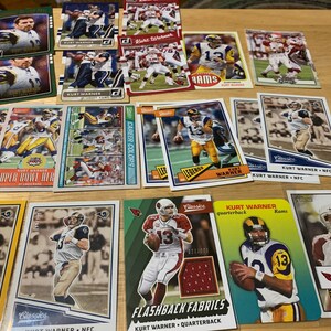 May include: A collection of Kurt Warner football trading cards featuring various images of the quarterback in different uniforms. The cards are from different sets, including "Super Bowl Heroes", "Legends", "Career Colors", "Classics", and "Flashback Fabrics".