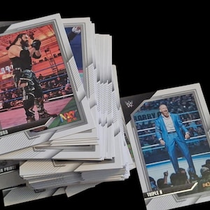 May include: A stack of wrestling trading cards featuring action shots of wrestlers in a wrestling ring. The cards have a white border and the WWE and NXT logos. The cards are on a black background.