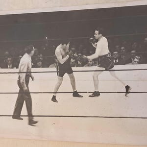 May include: Black and white photograph of a boxing match in progress. Two boxers in shorts and gloves are in the ring, with a referee observing. Spectators fill the background, watching the fight.