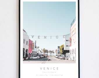 May include: Venice, California art print in a black frame. The image shows a street scene with buildings in pink, white, and other colors under a clear blue sky. The word "VENICE" is displayed across the street. Coordinates are also included.