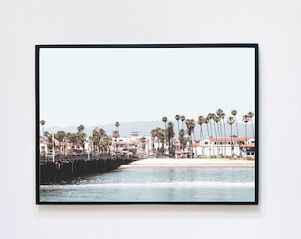May include: A framed photograph of a beach scene with a pier, palm trees, and buildings in the distance. The water is blue and the sky is light blue.