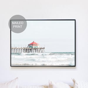 May include: A framed wall art print featuring a photograph of a pier extending into the ocean with a red-roofed building at the end and crashing waves in the foreground. A circular gray overlay in the top left corner contains the text MAILED PRINT.