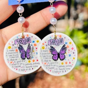 May include: A pair of white round earrings with a purple butterfly and glitter. The earrings have a gold chain and white pearl beads. The earrings have the text "SCRU" and a list of words on them.