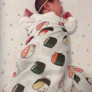 Op de afbeelding: Een baby gewikkeld in een witte deken met sushi-thema. De deken heeft kleurrijke illustraties van verschillende sushi-rollen. De baby draagt een witte onesie en een lichtblauwe strik-haarband. De achtergrond is een wit laken met roze sterren en manen.