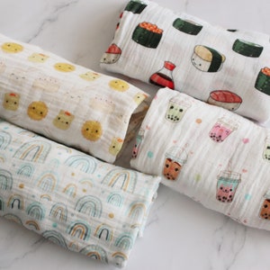 Op de afbeelding: Vijf witte muslin swaddles met verschillende schattige cartoonontwerpen. De ontwerpen omvatten sushi, boba-thee, regenbogen en dumplings.