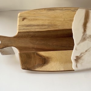 May include: A wooden cutting board with a white and brown epoxy resin inlay. The board has a handle and is perfect for serving cheese, charcuterie, or other appetizers.