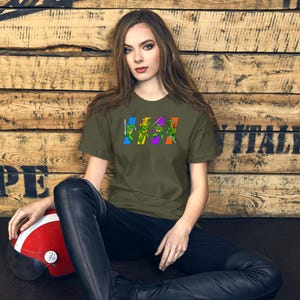 Könnte beinhalten: Ein olivgrünes T-Shirt mit einer Grafik der Teenage Mutant Ninja Turtles in einer Reihe, jede in einem andersfarbigen Rechteck. Die Schildkröten sind mit ihren Waffen dargestellt. Das T-Shirt wird von einer Person getragen.