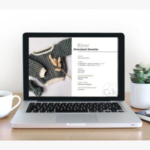 Op de afbeelding: Een laptop toont een breipatroon voor een oversized trui. Het patroon, getiteld "River", toont een grijs en cr&egrave;mekleurige gebreide trui. Het scherm toont ook garendetails, haaknaaldmaten en informatie over het vaardigheidsniveau. Een kopje en een plant staan op het bureau.