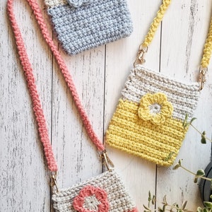 Pode incluir: Tr&ecirc;s bolsas de telefone de croch&ecirc; feitas &agrave; m&atilde;o em cores pastel: cinza e azul, cinza e amarelo e cinza e coral. Cada bolsa apresenta uma flor aplicada e uma al&ccedil;a tran&ccedil;ada combinando com fechos dourados. As bolsas s&atilde;o exibidas em uma superf&iacute;cie de madeira clara.