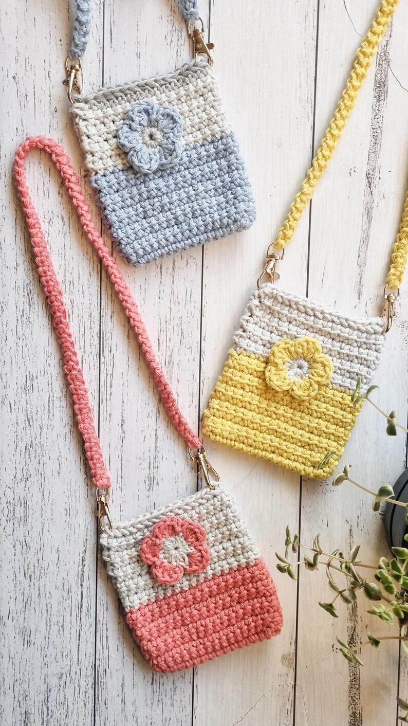 Pode incluir: Tr&ecirc;s bolsas de telefone de croch&ecirc; feitas &agrave; m&atilde;o em cores pastel: cinza e azul, cinza e amarelo e cinza e coral. Cada bolsa apresenta uma flor aplicada e uma al&ccedil;a tran&ccedil;ada combinando com fechos dourados. As bolsas s&atilde;o exibidas em uma superf&iacute;cie de madeira clara.