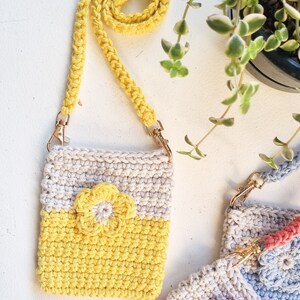 Pode incluir: Uma bolsa de ombro de croch&ecirc; feita &agrave; m&atilde;o em amarelo e creme com uma aplica&ccedil;&atilde;o de flor amarela. A bolsa tem uma al&ccedil;a amarela combinando com fechos de metal dourado. Uma planta com folhas verdes est&aacute; no fundo.