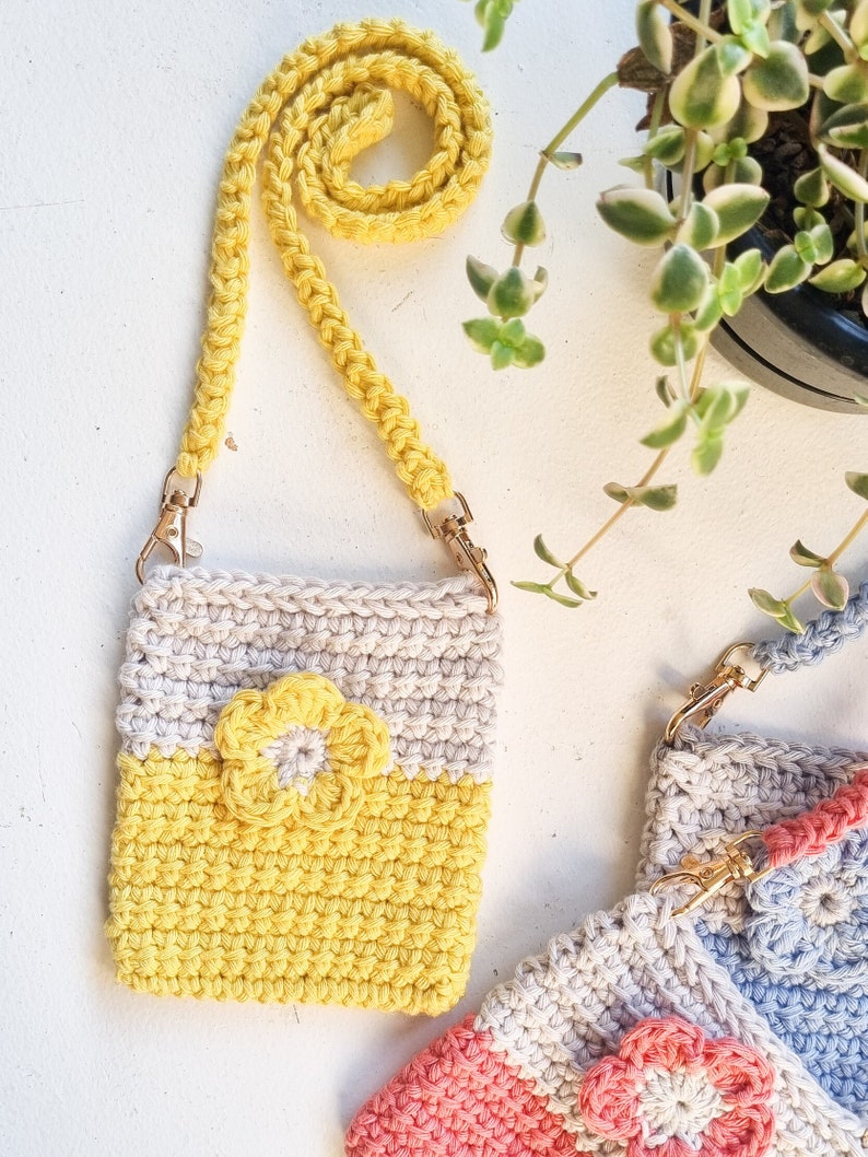 Pode incluir: Uma bolsa de ombro de croch&ecirc; feita &agrave; m&atilde;o em amarelo e creme com uma aplica&ccedil;&atilde;o de flor amarela. A bolsa tem uma al&ccedil;a amarela combinando com fechos de metal dourado. Uma planta com folhas verdes est&aacute; no fundo.