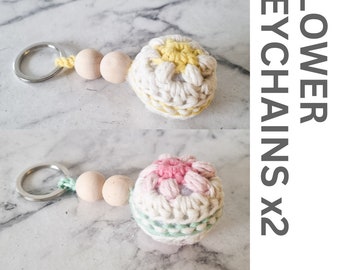 Könnte beinhalten: Ein Set aus zwei gehäkelten Blumen-Schlüsselanhängern mit Holzperlen. Die Schlüsselanhänger sind aus weißem Garn gefertigt und haben ein gelbes und pinkfarbenes Blumendesign. Der Text "FLOWER KEYCHAINS x2" und "8 PLY PDF PATTERN" ist im Bild sichtbar.
