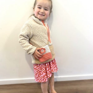Pode incluir: Uma pequena bolsa de croch&ecirc; coral e branca com um detalhe de flor &eacute; usada sobre uma jaqueta de l&atilde; bege e uma saia floral rosa. A bolsa tem uma al&ccedil;a longa e fina. A menina est&aacute; sorrindo.