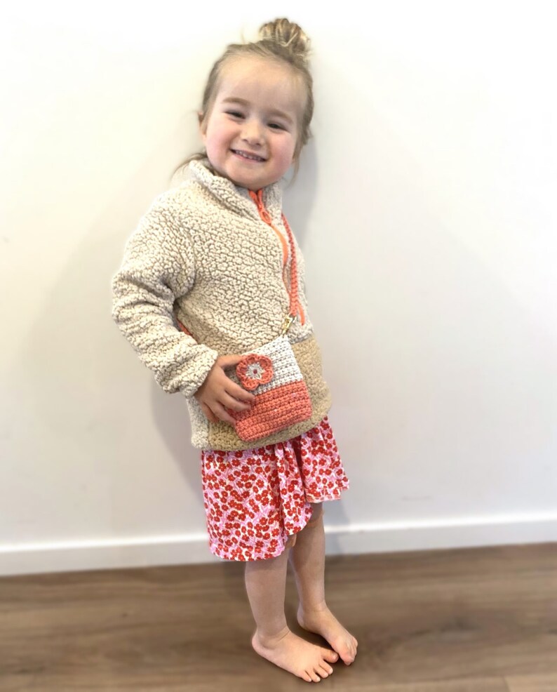 Pode incluir: Uma pequena bolsa de croch&ecirc; coral e branca com um detalhe de flor &eacute; usada sobre uma jaqueta de l&atilde; bege e uma saia floral rosa. A bolsa tem uma al&ccedil;a longa e fina. A menina est&aacute; sorrindo.