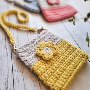 Pode incluir: Uma bolsa de ombro de croch&ecirc; feita &agrave; m&atilde;o em amarelo e cinza, com um detalhe de flor amarela. A bolsa tem uma al&ccedil;a tran&ccedil;ada amarela e ferragens douradas. Outras bolsas de croch&ecirc; em rosa e azul est&atilde;o ligeiramente desfocadas no fundo.