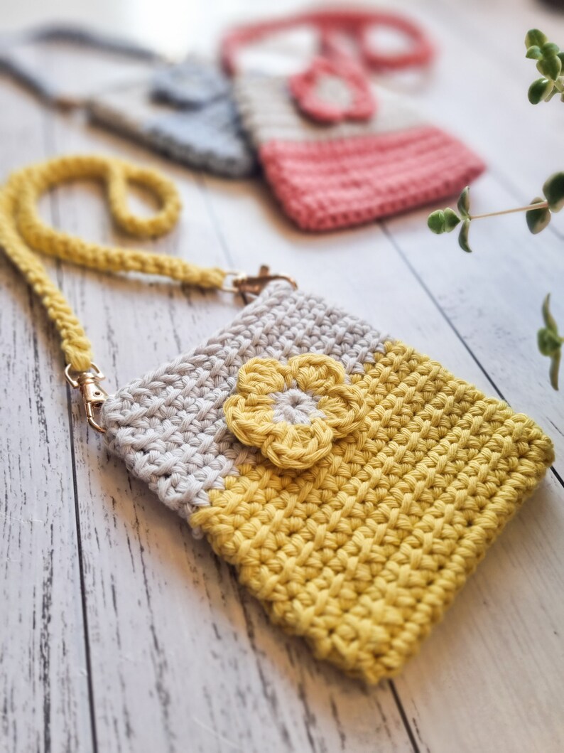 Pode incluir: Uma bolsa de ombro de croch&ecirc; feita &agrave; m&atilde;o em amarelo e cinza, com um detalhe de flor amarela. A bolsa tem uma al&ccedil;a tran&ccedil;ada amarela e ferragens douradas. Outras bolsas de croch&ecirc; em rosa e azul est&atilde;o ligeiramente desfocadas no fundo.