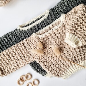 Op de afbeelding: Drie handgebreide truien in grijs, beige en cr&egrave;me, met een textuursteekpatroon. De truien zijn gerangschikt op een wit oppervlak, met een bol wol en houten ringen in de buurt. De truien hebben een ronde hals en lange mouwen.