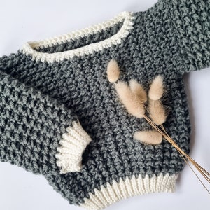 Op de afbeelding: Een grijze gehaakte trui met witte afwerking op de halslijn, manchetten en zoom. De trui heeft een gestructureerd steekpatroon en lange mouwen. Gedroogde beige bloemen zijn bij de trui geplaatst.