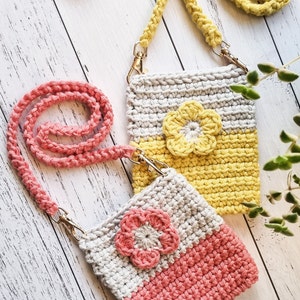 Pode incluir: Duas bolsas de telefone de croch&ecirc; feitas &agrave; m&atilde;o com al&ccedil;as transversais. Uma bolsa &eacute; cinza e amarela com uma flor amarela, a outra &eacute; cinza e coral com uma flor coral. Ambas t&ecirc;m fechos dourados. Uma terceira bolsa azul &eacute; vis&iacute;vel ao fundo.