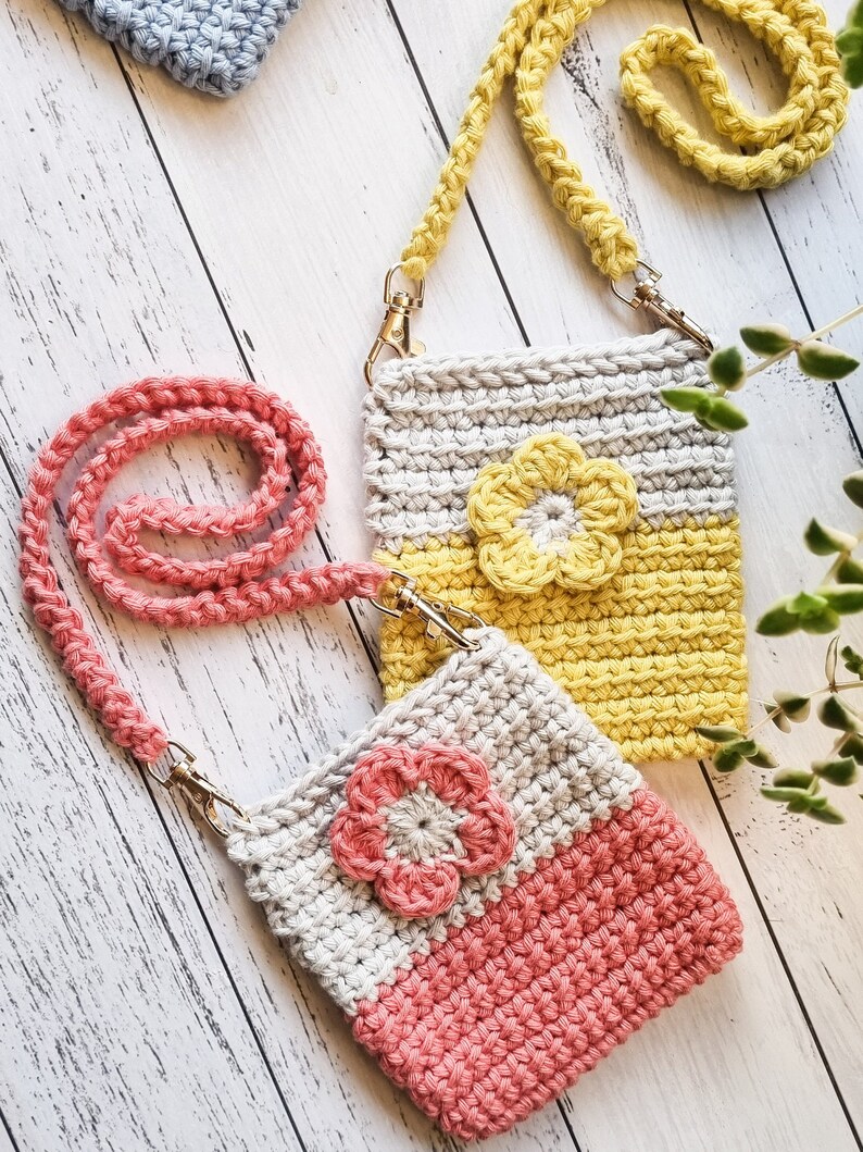 Pode incluir: Duas bolsas de telefone de croch&ecirc; feitas &agrave; m&atilde;o com al&ccedil;as transversais. Uma bolsa &eacute; cinza e amarela com uma flor amarela, a outra &eacute; cinza e coral com uma flor coral. Ambas t&ecirc;m fechos dourados. Uma terceira bolsa azul &eacute; vis&iacute;vel ao fundo.