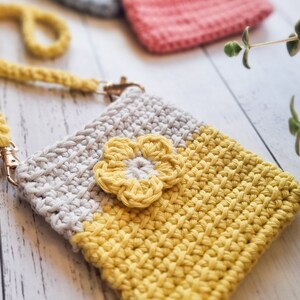 Pode incluir: Capa de telefone de croch&ecirc; feita &agrave; m&atilde;o em amarelo e bege com um detalhe floral amarelo. A capa tem uma al&ccedil;a de pulso e um fecho dourado. Outras capas de croch&ecirc; em rosa e azul s&atilde;o vis&iacute;veis no fundo.